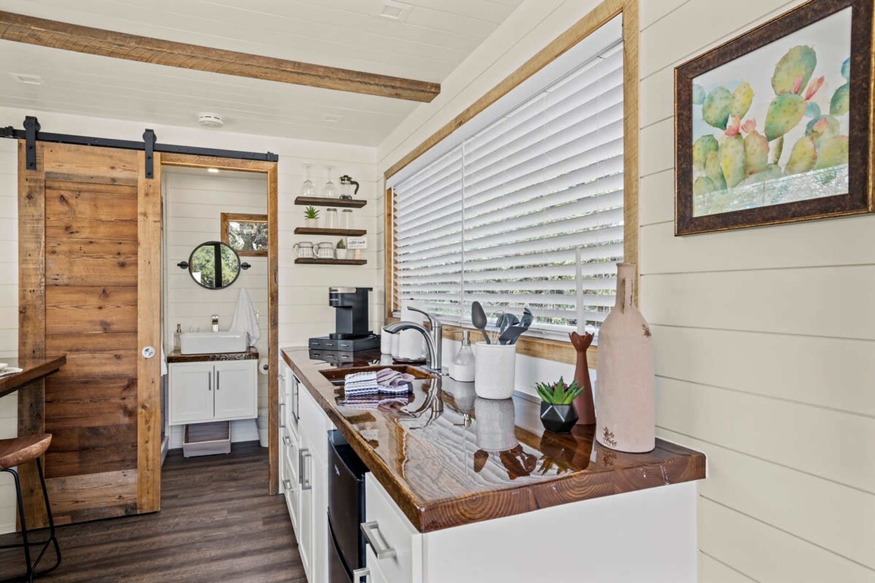 A kitchen with a wooden sliding door, white cabinets, a coffee machine, and a window with blinds.
