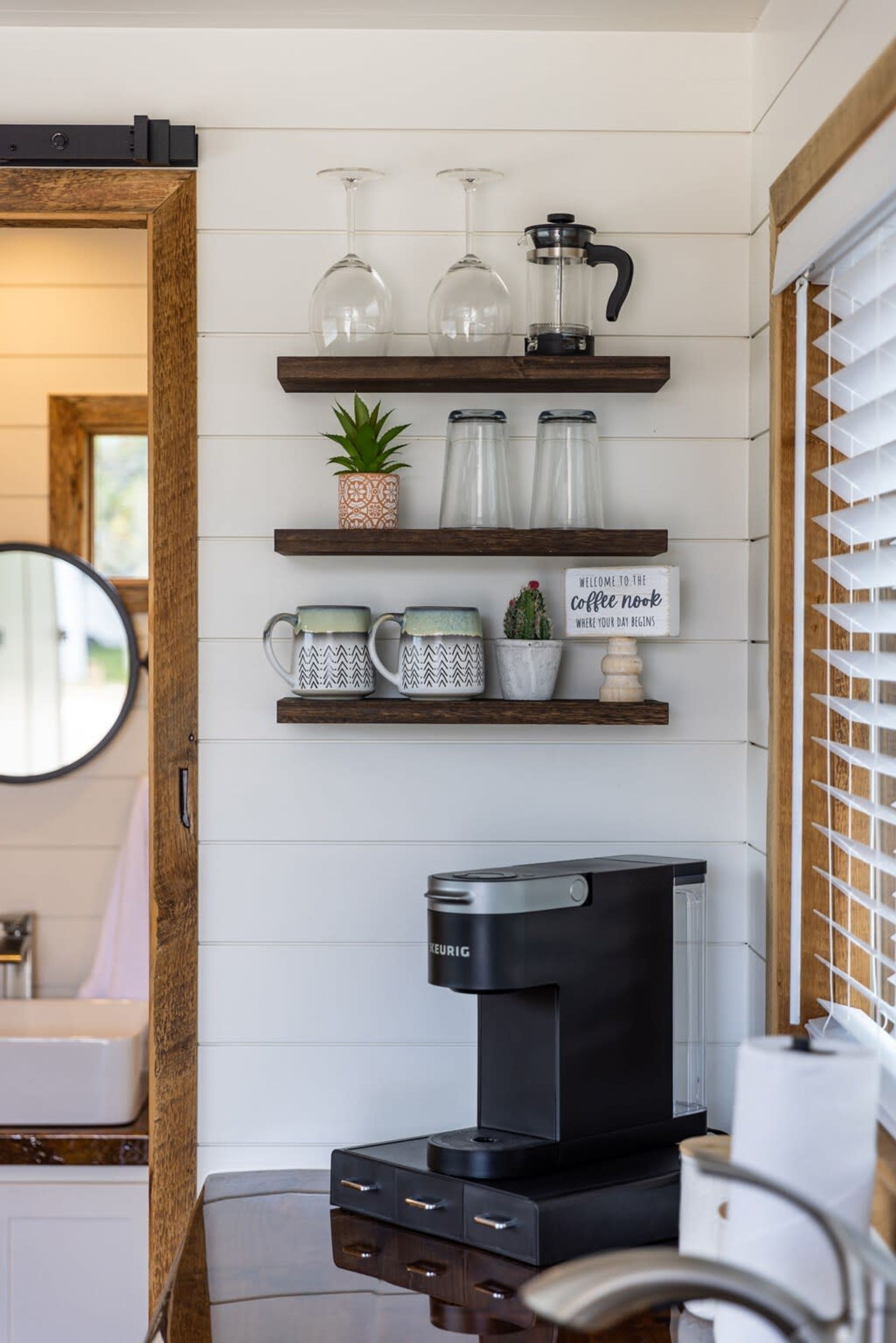 A kitchen with a coffee maker, shelves with mugs, a sign, and a potted plant.