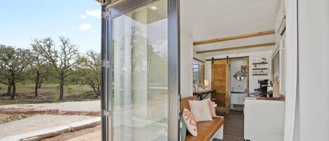 A modern interior with a sliding glass door leading to a deck, a cozy seating area with cushions, and a kitchen with white cabinets and wooden accents.