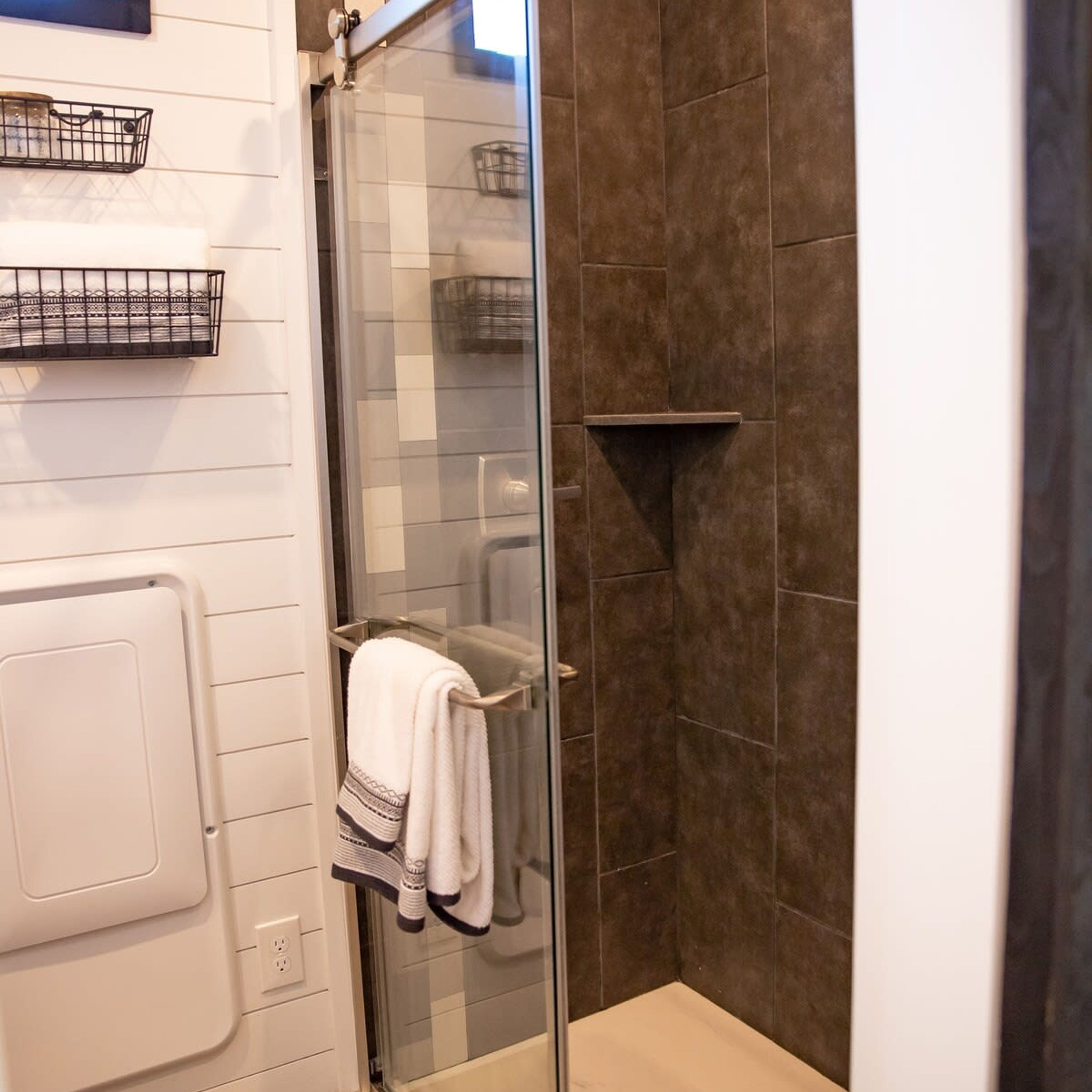 Un baño con cabina de ducha de vidrio, un perchero con toalla y dos cestas fijadas a la pared.