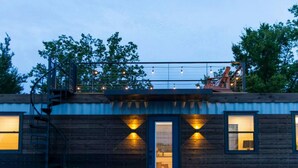 A rustic cabin-style building with a dark wooden exterior, a blue door, and two windows with warm lighting. - New Luxury Shipping Container (Waco)