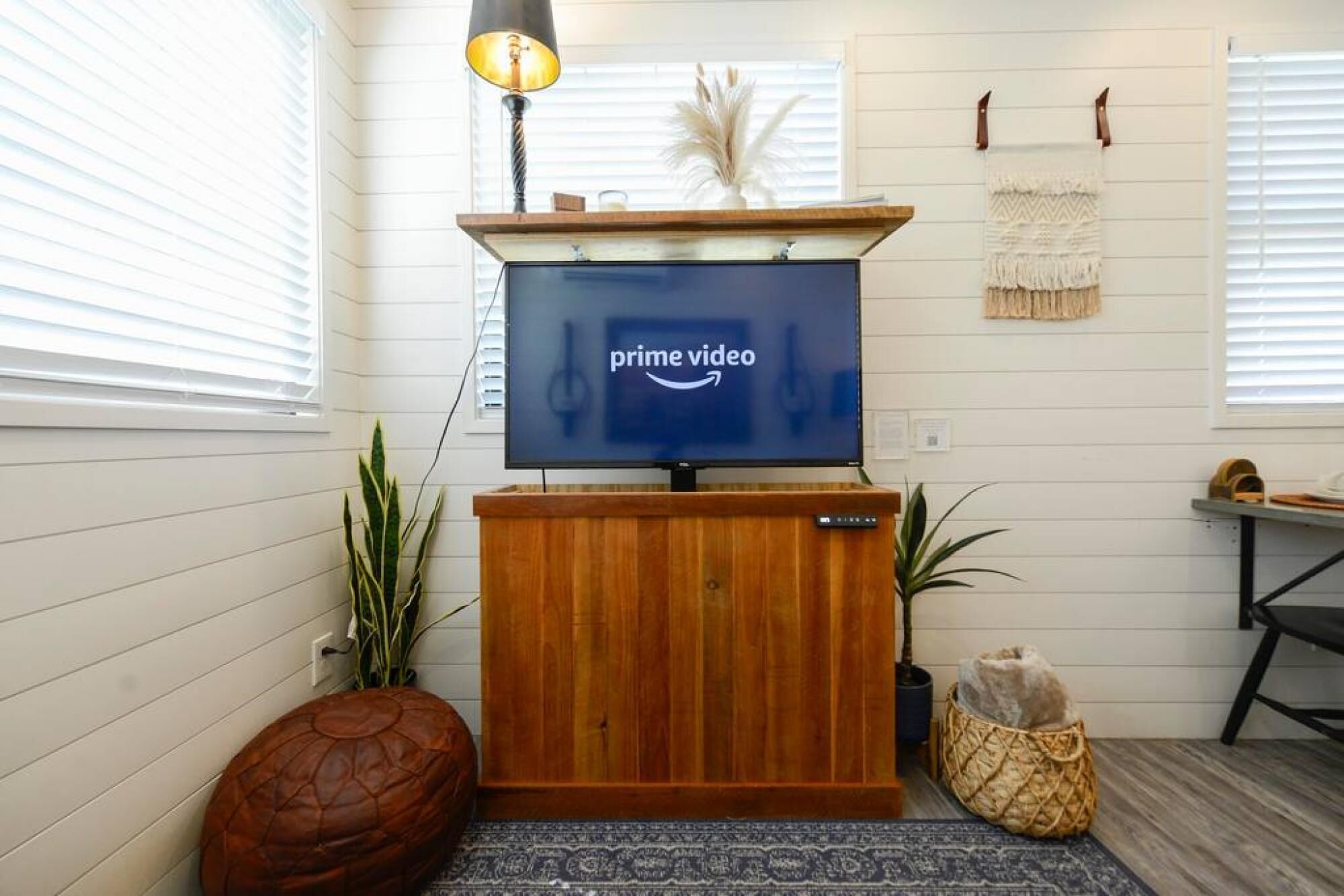 A living room with a flat-screen TV displaying the Prime Video logo, a wooden entertainment unit, a brown leather ottoman, a patterned rug, and a small plant.