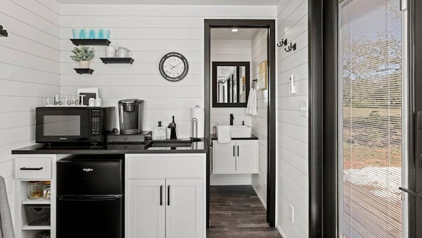 A modern kitchen with white cabinetry, a black refrigerator, a microwave, and a coffee maker. There is a clock on the wall and a window with blinds. - New Wilderness Hideout-cozy Container Home (Fredericksburg)