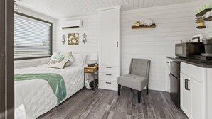 A compact, well-lit room with a bed, a small seating area, a kitchenette, and a window with blinds. - New Wilderness Hideout-cozy Container Home (Fredericksburg)