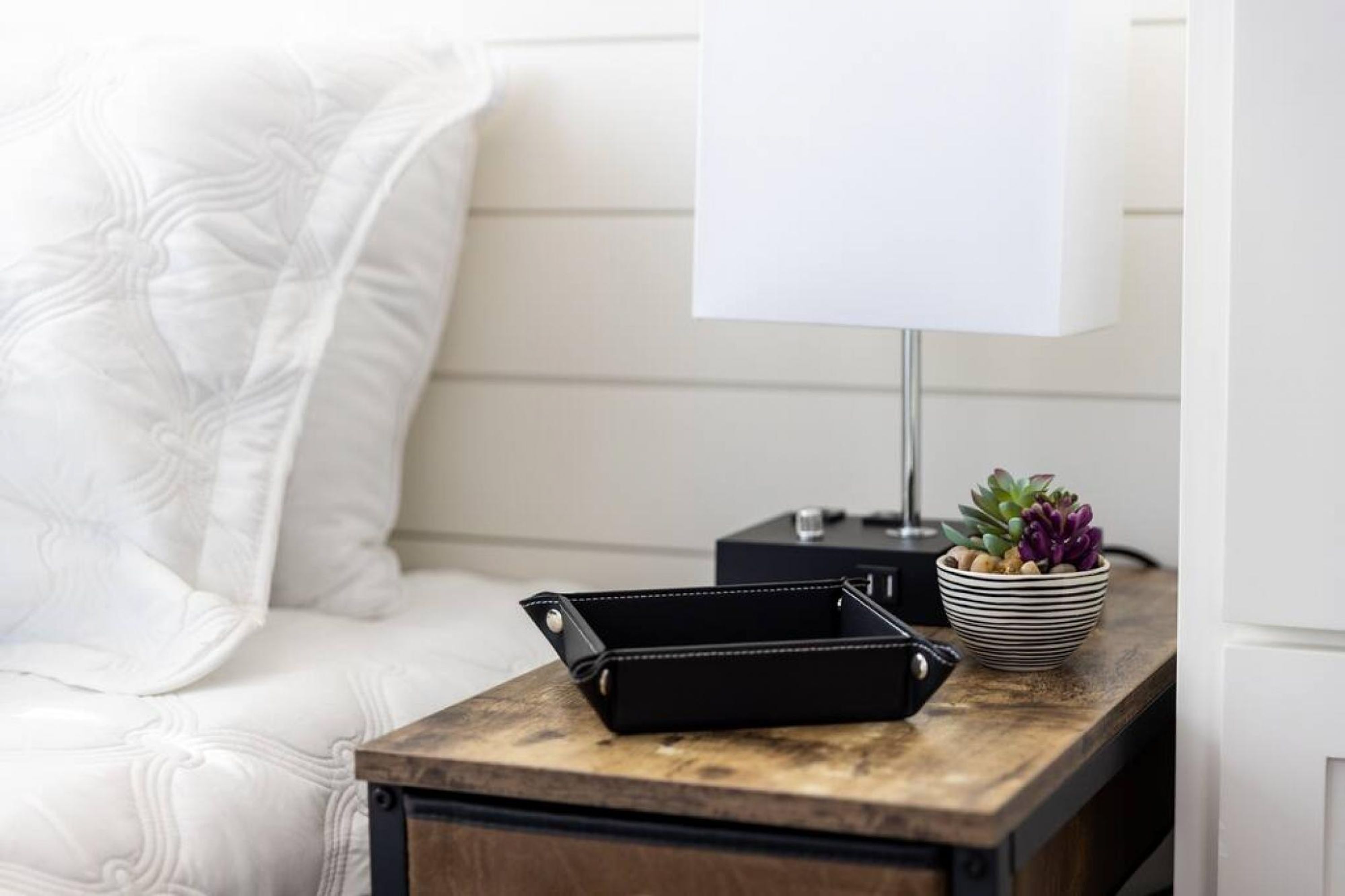 Ein ordentlich bezogenes Bett mit einem weißen Kopfkissen, ein schwarzer Nachttisch mit einer Lampe, eine schwarze Brieftasche und eine gestreifte Schale mit Sukkulenten.