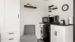 A compact kitchen with white cabinetry, a grey upholstered chair, a microwave, a coffee maker, and a refrigerator. - New Wilderness Hideout-cozy Container Home (Fredericksburg)