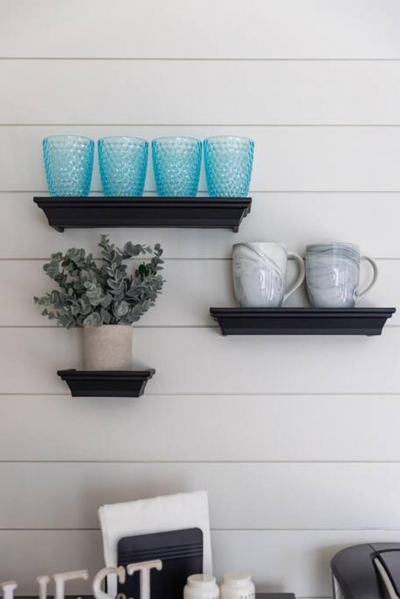 Three black shelves against a white wall, each holding different items: a potted plant, blue glass cups, and white ceramic mugs.