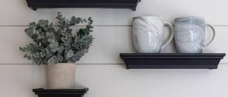 Three black shelves against a white wall, each holding different items: a potted plant, blue glass cups, and white ceramic mugs.