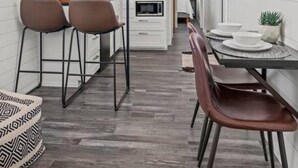 Een smalle keuken met witte muren, houten balken en een klok. Er staan twee barkrukken en een eettafel met stoelen.