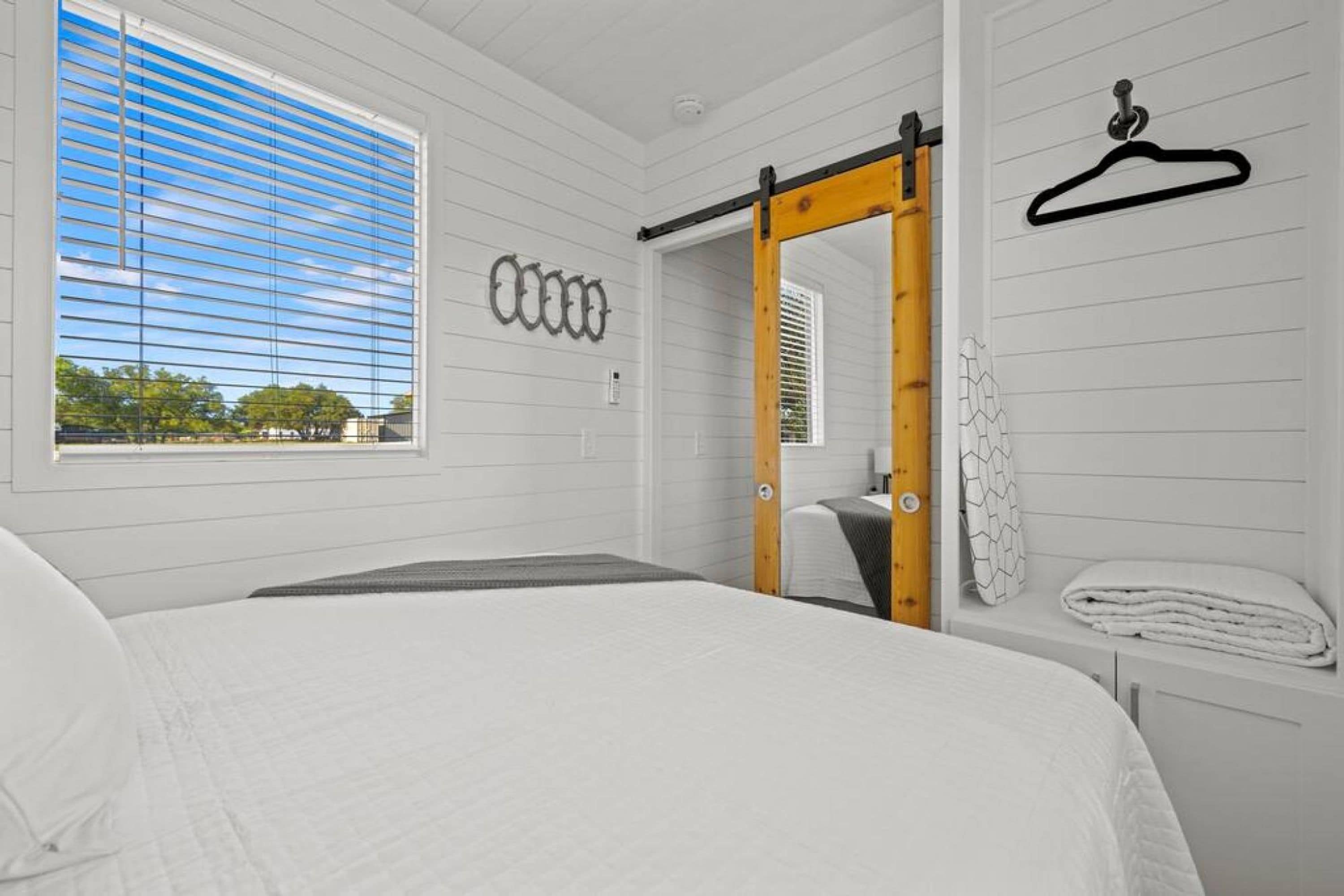 Une chambre avec un grand lit, une fenêtre avec des stores, une porte en bois et un porte-manteau fixé au mur.