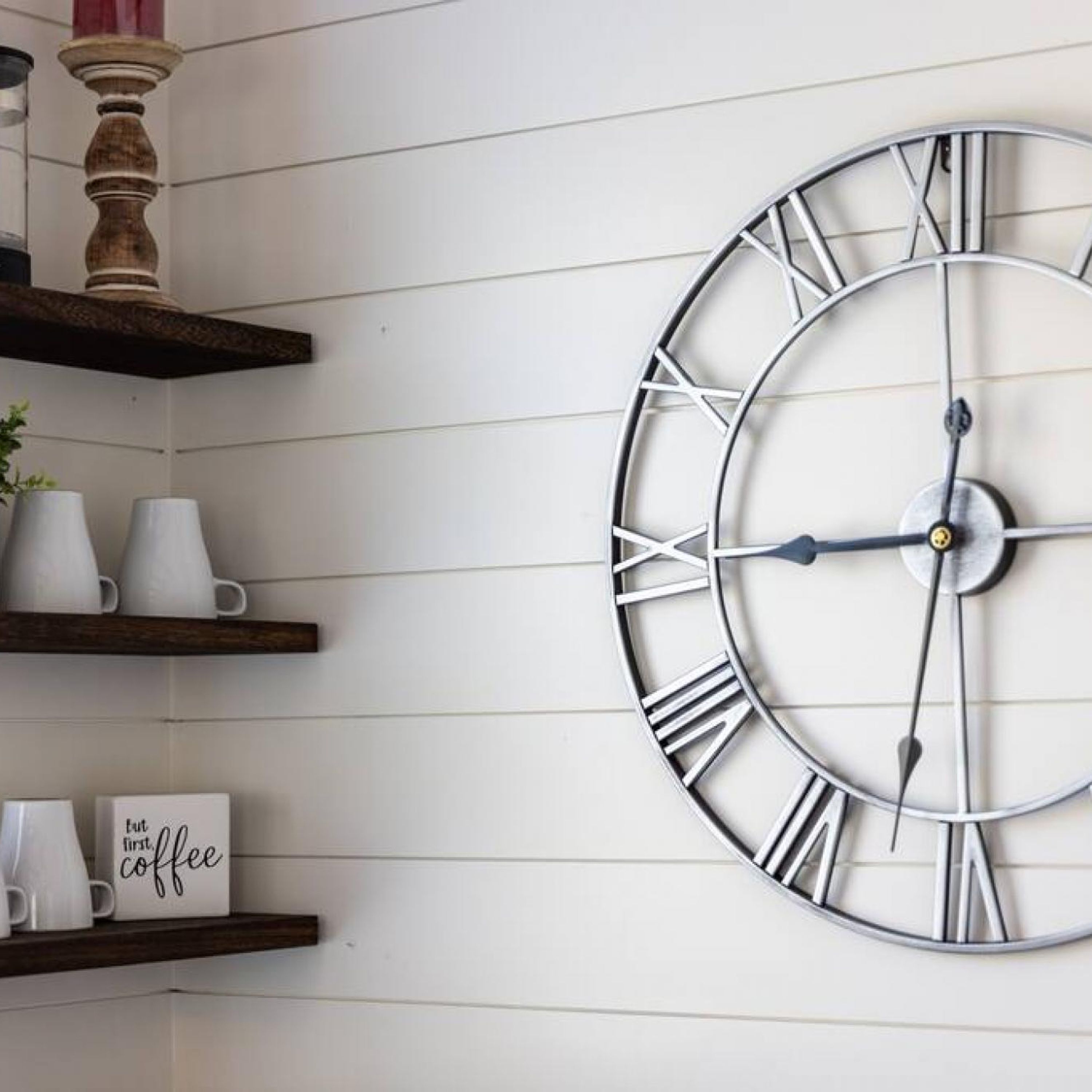 A wall-mounted clock with Roman numerals, shelves with white mugs, and a sign that says "but first coffee".
