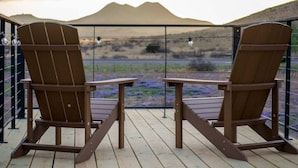 Dos sillas Adirondack de madera en una terraza de madera con vistas a montañas y flores púrpuras.