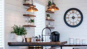 Una cocina con lámparas colgantes, un reloj, estantes con plantas y una cafetera.