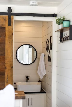 A bathroom with a white sink, a round mirror, and a towel hanging on the wall. - New The Wildflower Cozy Container Home (Fredericksburg)