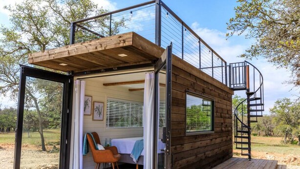 A modern cabin with a glass door entrance, a wooden deck, and a spiral staircase. - New The Wildflower Cozy Container Home (Fredericksburg)