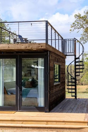 A modern cabin with a spiral staircase, large sliding glass doors, and a deck. - New The Wildflower Cozy Container Home (Fredericksburg)