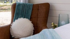 A room with a brown leather chair, a teal throw blanket, and a white textured pillow. There are two framed botanical prints on the wall. - New The Wildflower Cozy Container Home (Fredericksburg)