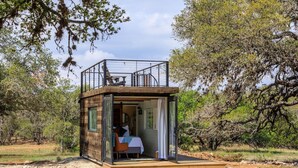 A small, open-air cabin with a bed and a table, surrounded by trees. - New The Wildflower Cozy Container Home (Fredericksburg)