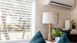 A modern living room with a large window, a wooden floor lamp, and a potted plant. - New The Wildflower Cozy Container Home (Fredericksburg)