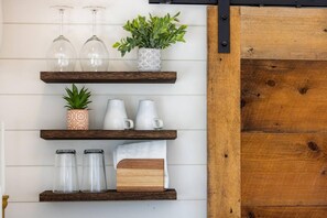 A wall-mounted shelf with various items including wine glasses, a potted plant, pitchers, and a candle holder. - New The Sunrise Cozy Container Home (Fredericksburg)