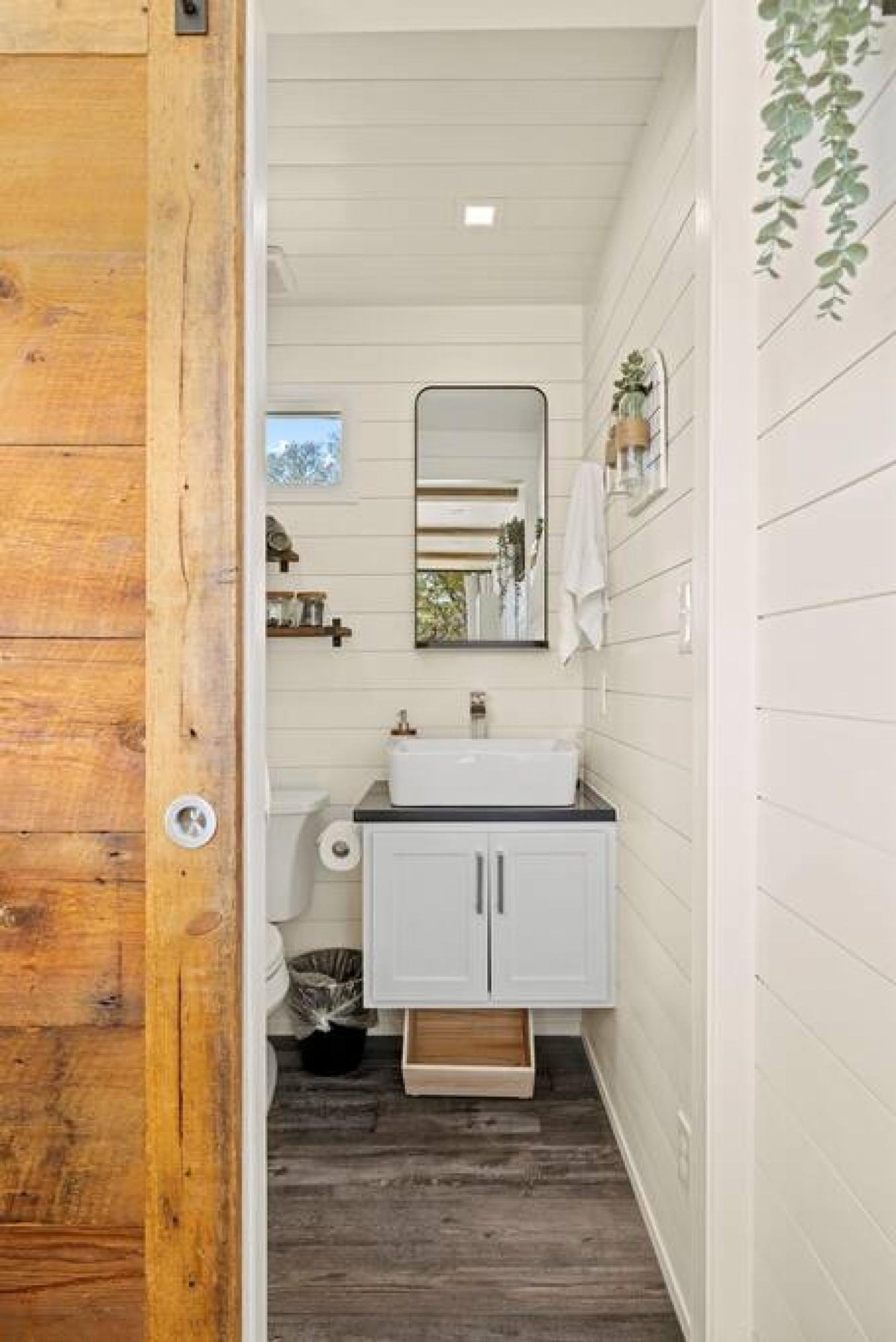 A narrow bathroom with a wooden door, white walls, a white sink, and a mirror.