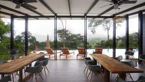 Un vaste espace dédié à la salle à manger, avec une longue table en bois et des chaises, de grandes fenêtres donnant sur la forêt et des ventilateurs de plafond.