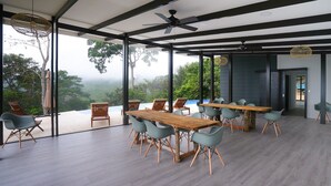 Un vaste espace dédié à la salle à manger, avec une longue table en bois et des chaises modernes, ainsi que de grandes fenêtres offrant une vue sur une végétation luxuriante.