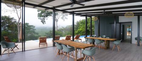 A spacious dining area with a long wooden table and modern chairs, large windows offering a view of lush greenery.