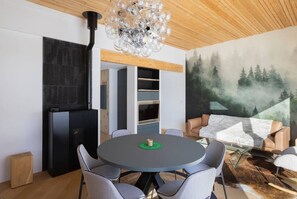 Un salon moderne avec une table ronde, des fauteuils gris, une cheminée noire et une fresque représentant une forêt sur le mur.