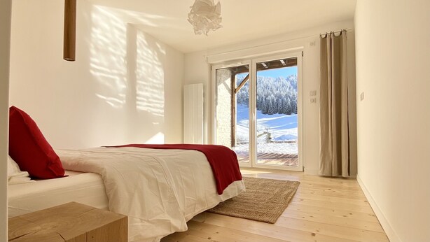 Un dormitorio con cama, un banco de madera, un ventanal con vistas a un paisaje nevado y una lámpara de techo.