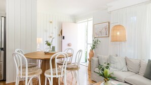 A dining area with a round wooden table and white chairs, a sofa with striped cushions, and a wicker lamp.