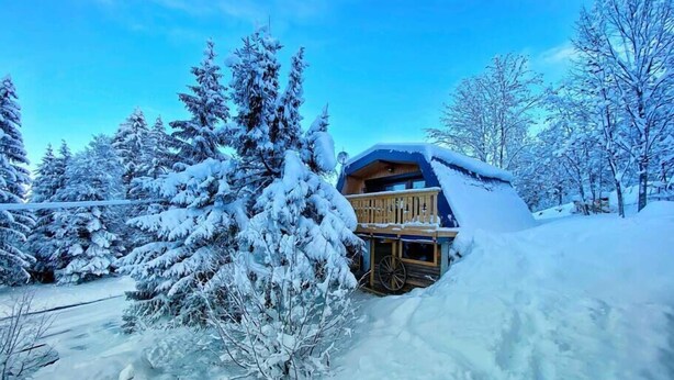 Un chalet enneigé doté d’une terrasse en bois et d’une grande fenêtre, entouré d’arbres chargés de neige.