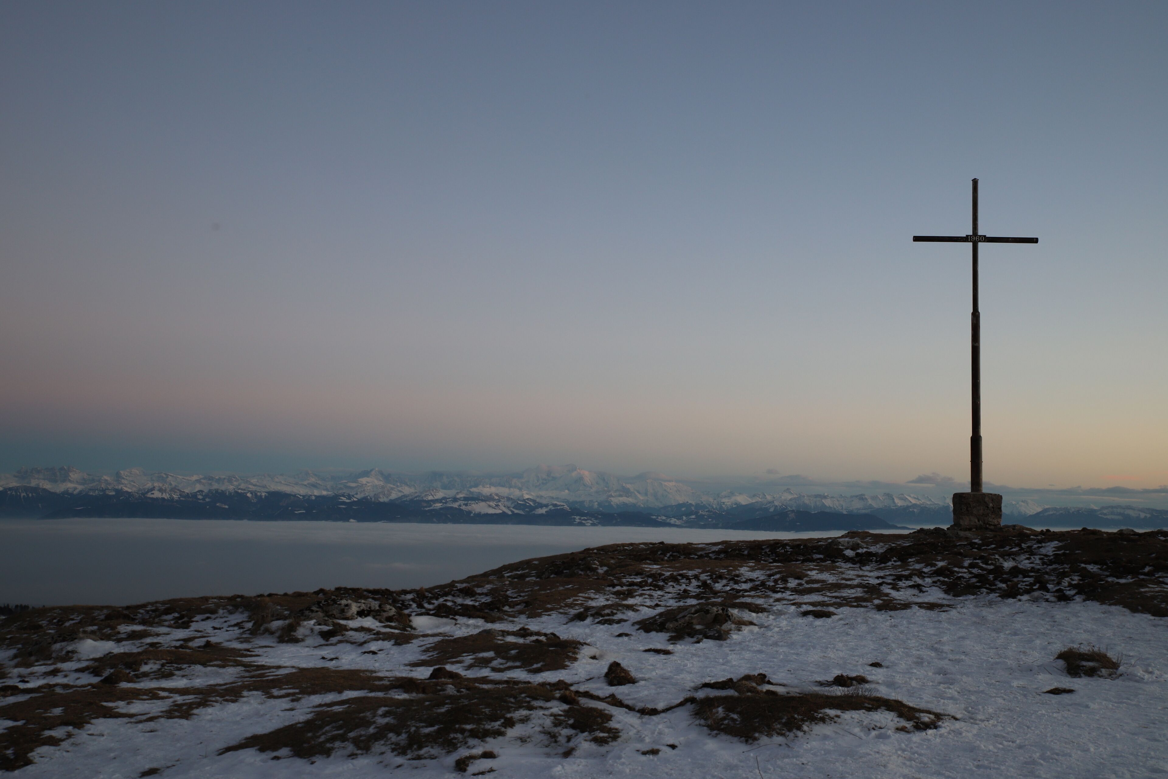 Uma cruz em um afloramento rochoso com vista para montanhas cobertas de neve ao fundo.