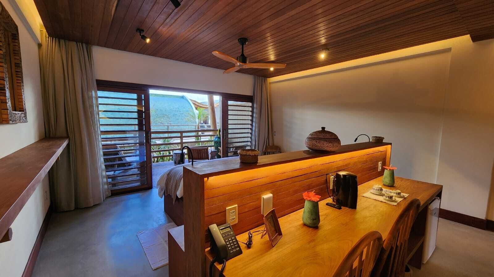 A hotel room with a wooden reception desk, a dining area with a table and chairs, a bed, and a large window with a view of the outdoors.