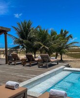 Zona junto a la piscina con sillones de descanso y una pequeña piscina, rodeada de palmeras y un cielo azul despejado.