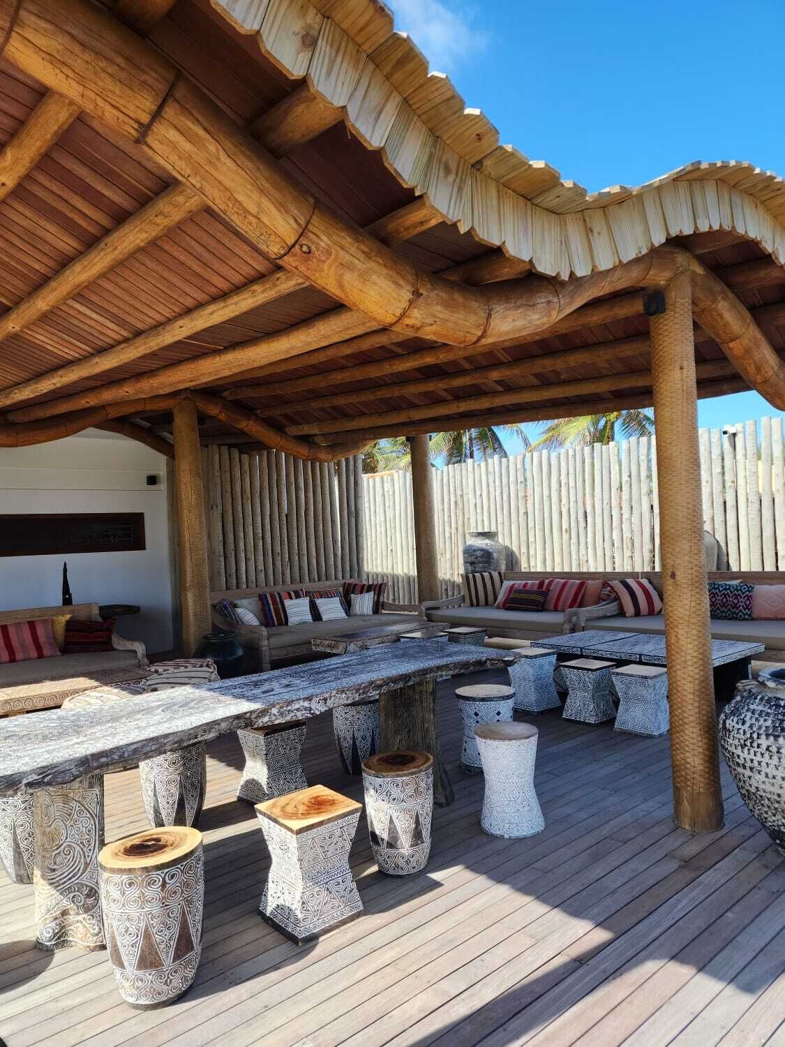 En udendørs terrasse med træterrasse, et overdækket opholdsområde med hynder og et stort bord med mønstrede bænke.