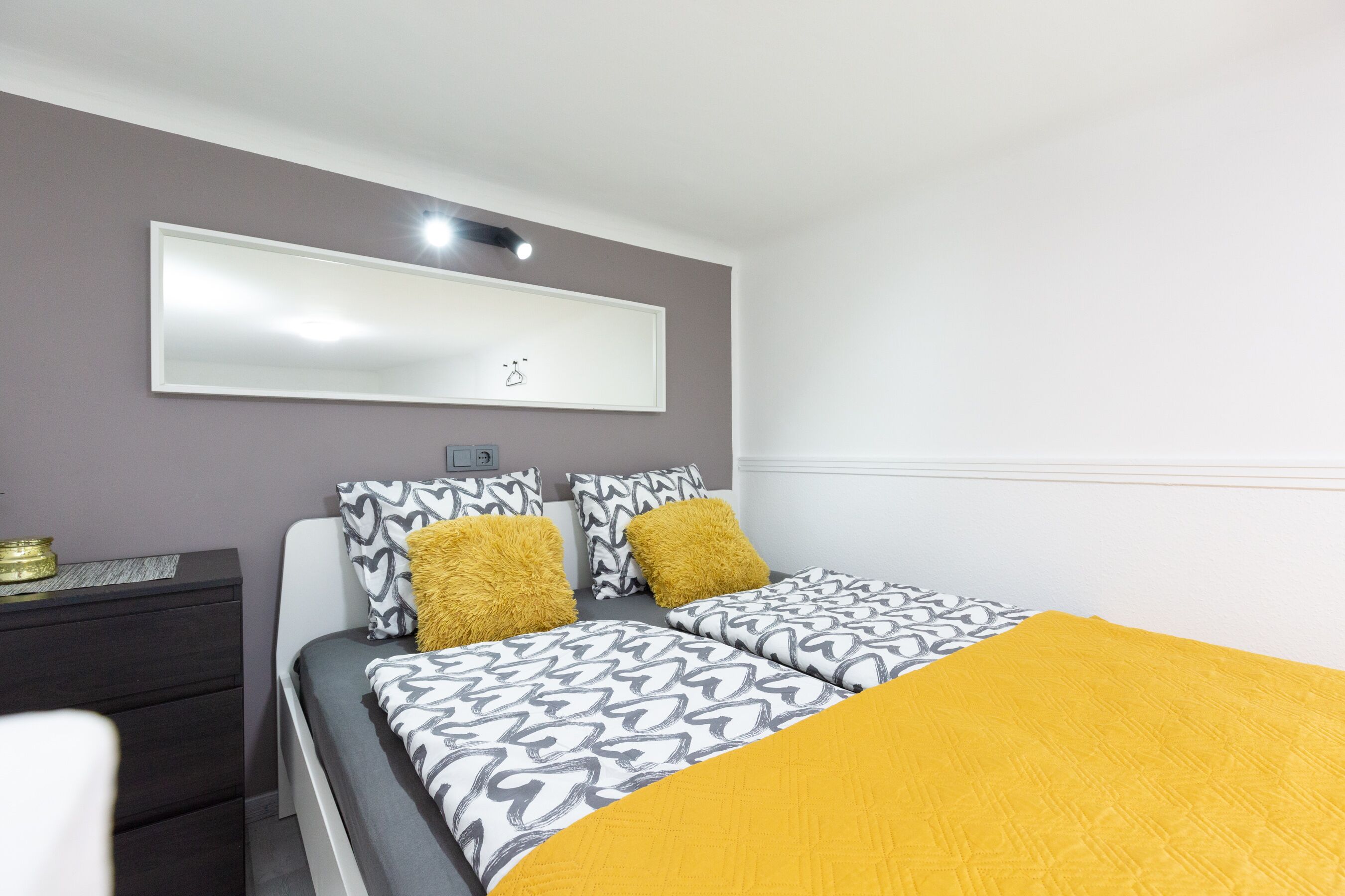 Een slaapkamer met een bed, twee kussens, een nachtkastje en een spiegel aan de muur.
