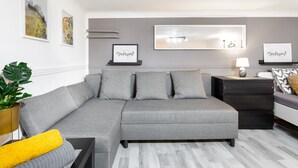 Un salon moderne avec un canapé d’angle gris, une petite table d’appoint, une plante en pot et un panneau encadré sur lequel on peut lire « Budapest ».