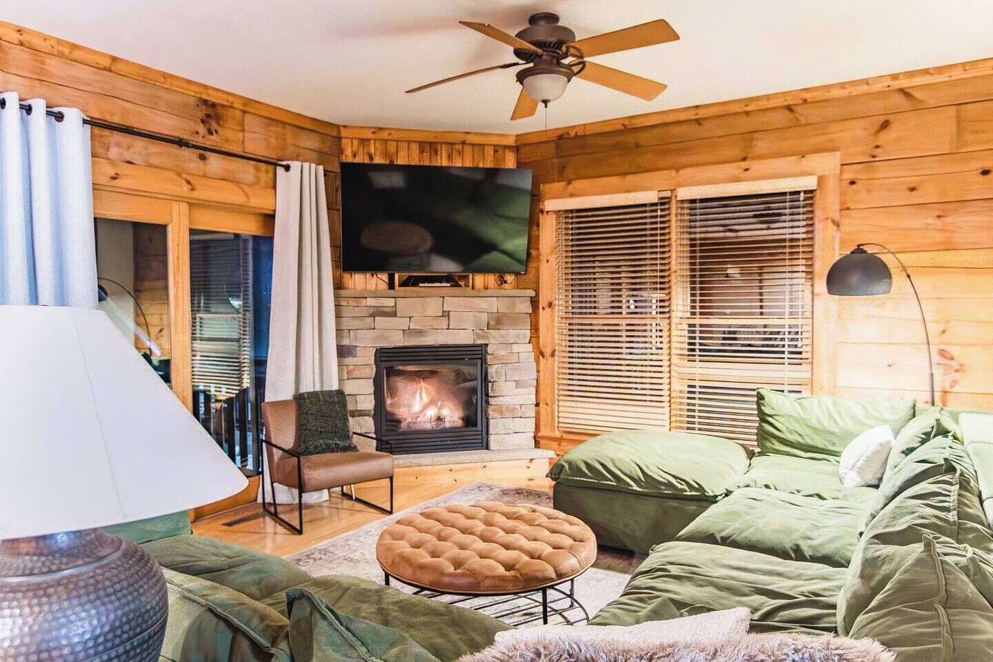 A cozy living room with a fireplace, wooden walls, a ceiling fan, and a green sofa.
