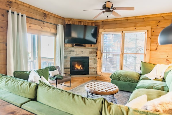 A cozy living room with a stone fireplace, green sofas, a flat-screen TV, and a ceiling fan. - Great for Families! Private Hot Tub, Views, Walk to Slopes (Snowshoe)