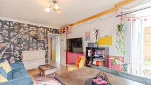 A cozy living room with a blue sofa, wooden coffee table, and a TV. There are shelves with books and decorative items, and a door leading to another room. - Vibrant 1BD Flat - 8 Mins to Dalston, Hackney! (London)