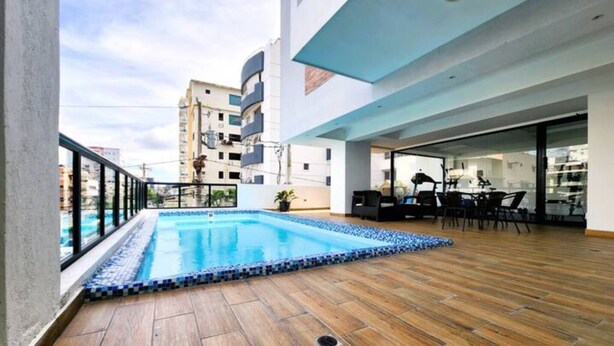 Un'area piscina sul tetto con un pontile in legno, una zona relax con sedie e un tavolo, e vista sullo skyline urbano.