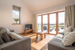 A modern living room with a sofa, a TV on a wooden stand, and a table. Large windows offer a view of the outdoors. - Hafod-Llecheiddior | A self catering cottage that sleeps 5 guests in 3 bedrooms (Bryncir)