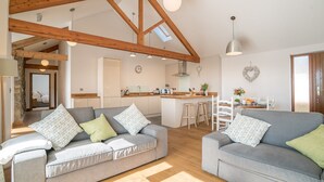 Un vaste espace de vie avec deux canapés gris, un plafond en bois et une cuisine à l’arrière-plan.