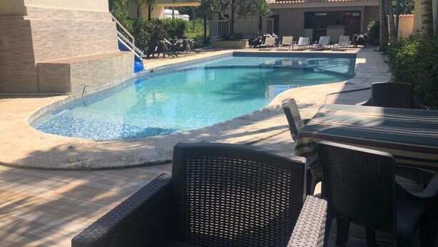 A swimming pool with lounge chairs and a table, surrounded by palm trees and a building with a tiled roof. - Hotel Cambri (Nagua)