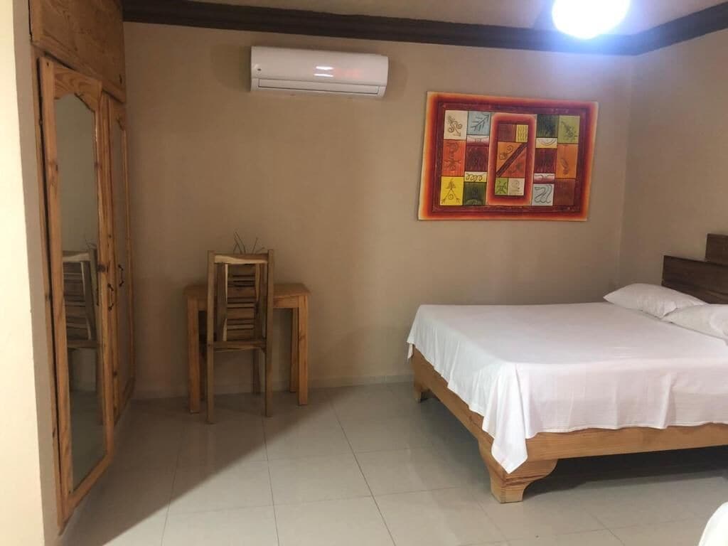 Une chambre à coucher avec un lit, un petit tabouret en bois, un climatiseur fixé au mur et une œuvre d’art aux couleurs vives.