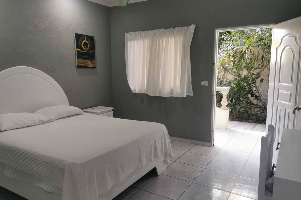 A bedroom with a bed, a nightstand, a window with curtains, and a door leading to an outdoor area.