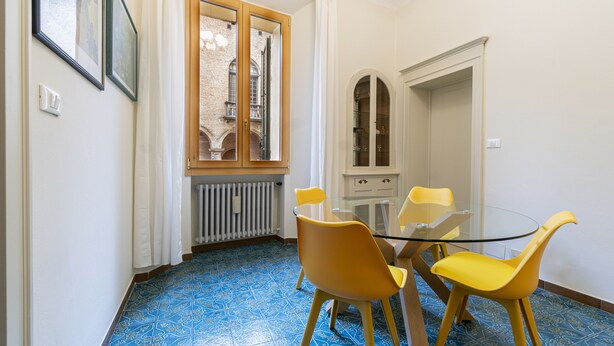 Ein Essbereich mit Glastisch und gelben Stühlen, einem Heizkörper unter dem Fenster und einem weißen Schrank mit Glastüren.