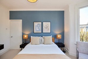 A bedroom with a bed, two bedside tables, two wall-mounted lamps, and two framed artworks on the wall.