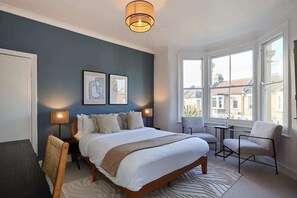 Une chambre à coucher avec un grand lit, deux tables de chevet, une chaise et une fenêtre donnant sur des immeubles.